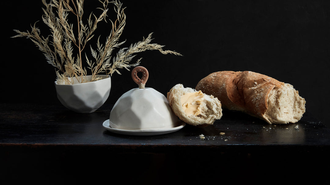 DBO HOME battuto collection featuring white faceted butter dish with leather handle and white faceted bowl with buttered bread loaf on a black background