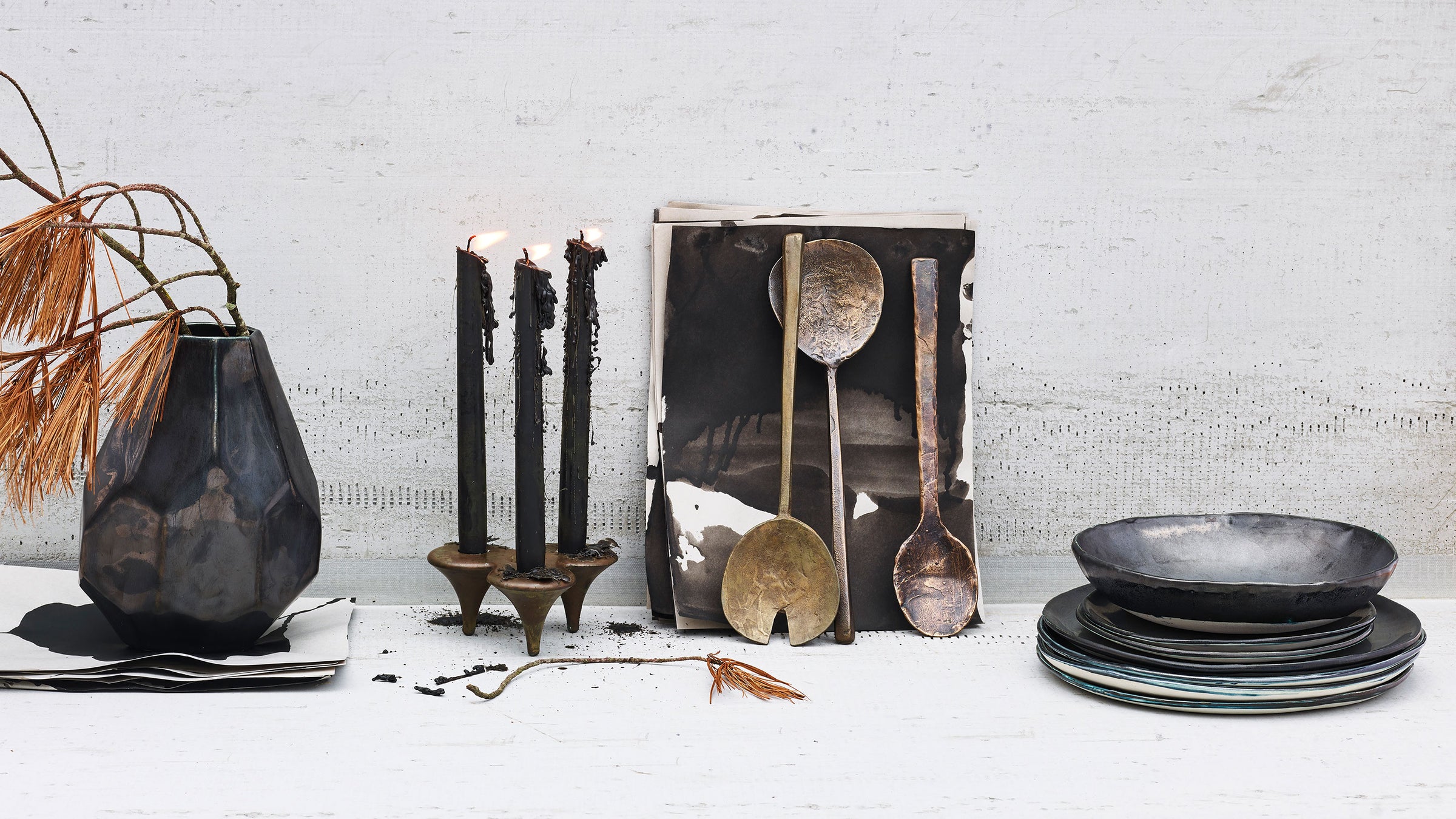 Artisan handcast bronze candlesticks, handcast bronze serveware and hand cast bronze spoon with black candles and black ceramic plates and vase on concrete background with pine needles