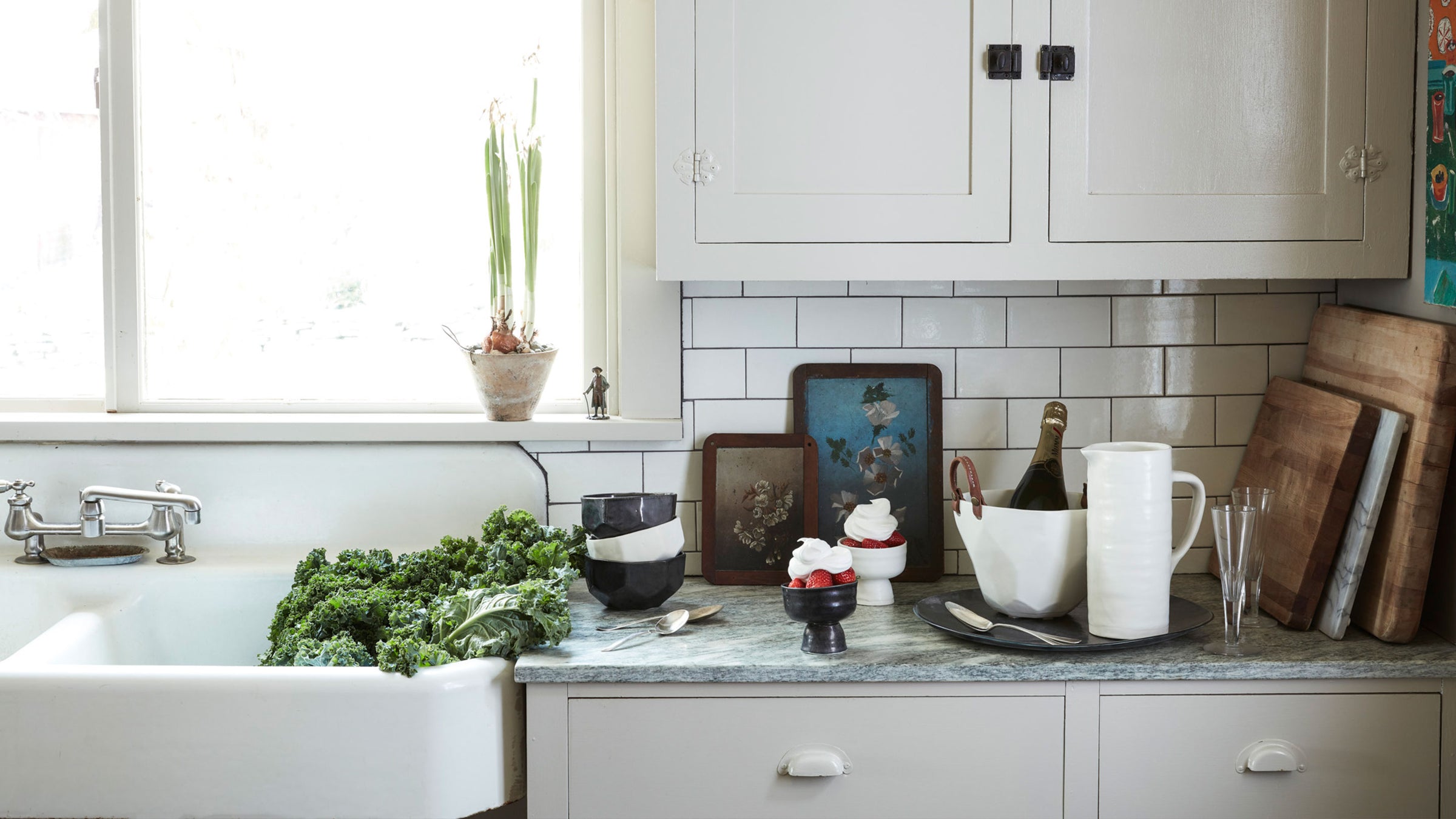 DBO HOME black and white artisan ceramic vessels, pitcher, serving bowls on a kitchen counter with subway tile and farm sink with kale