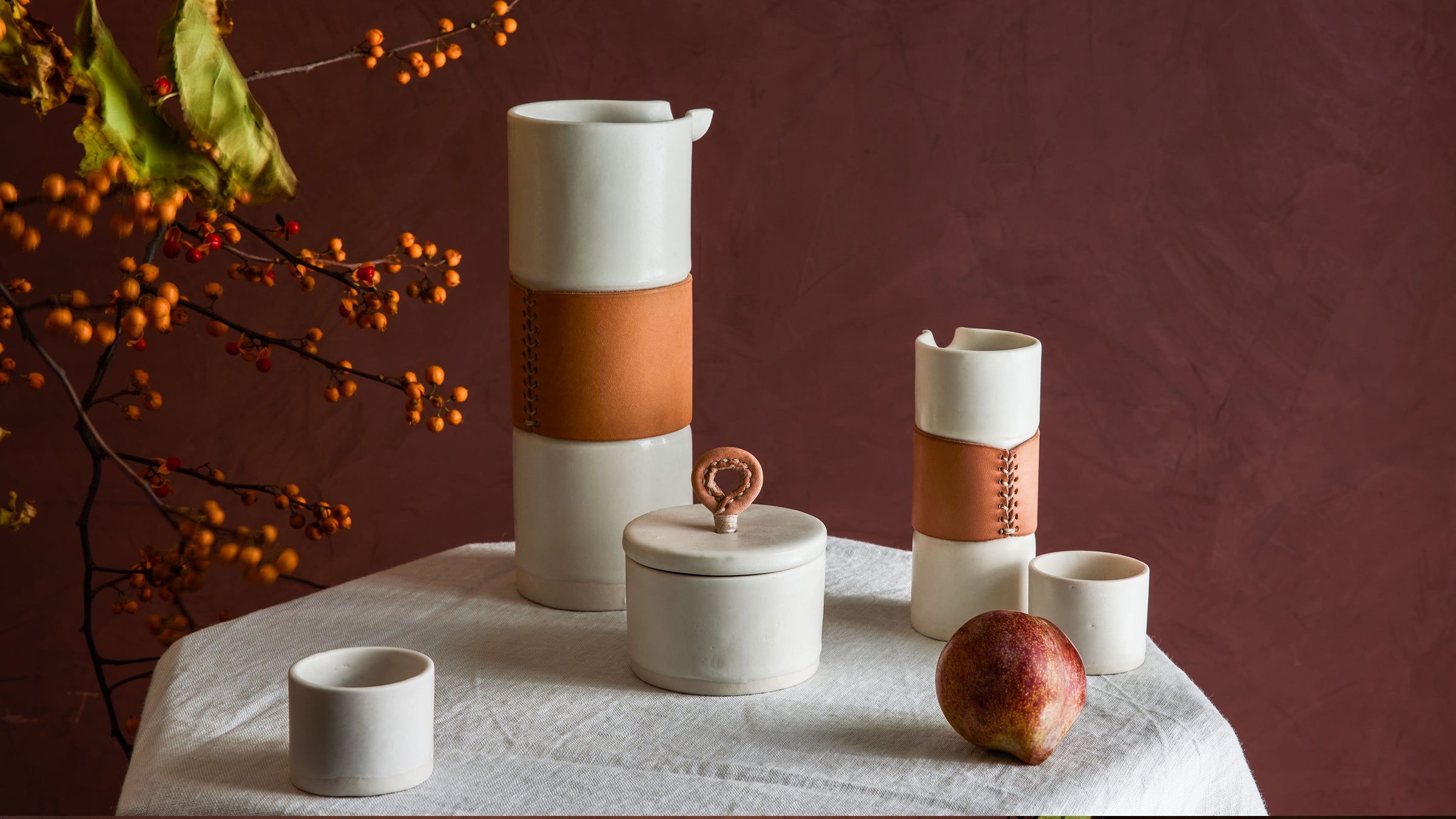 dbo home luxury handmade white porcelain and leather pitchers, salt cellar, and sake cups on linen table cloth with winter berries and fruit