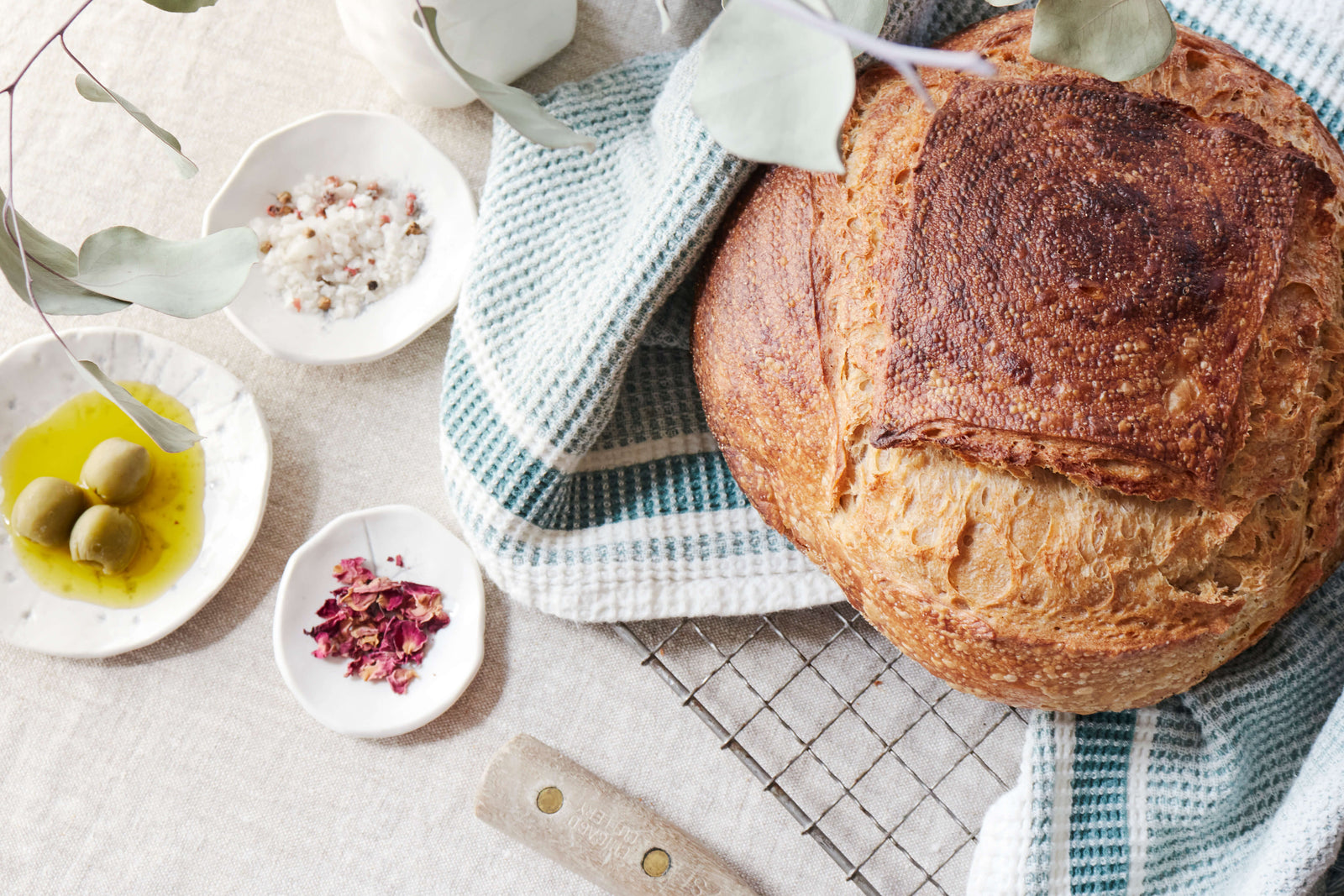 A loaf of crusty sourdough bread from Bourke Street Bakery on a blue and white waffle weave tea towel with salt, herbs, oil and olives in vintage DBO dipping bowls