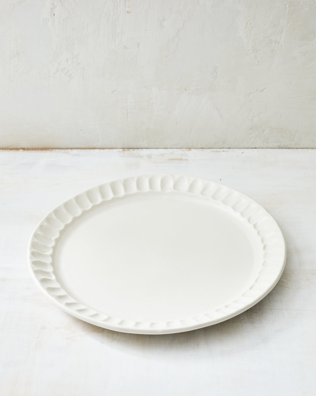 DBO HOME Nonna Dinner Plate in porcelain with a shimmering white satin glaze, revealing the natural warmth of the clay beneath. The softly fluted rim and gentle curves echo vintage European forms, while remaining modern and minimal. Photographed on a neutral-toned surface in soft, natural light.