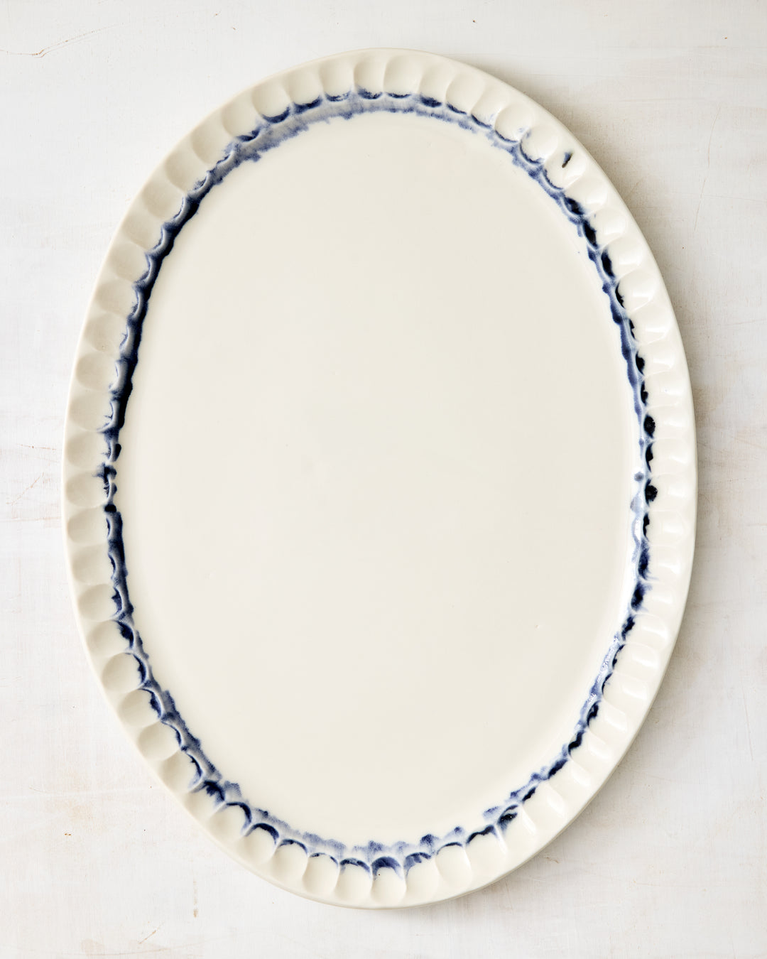 DBO HOME Nonna Oval Platter in Indigo, featuring a hand-painted cobalt rim over glossy clear glaze on white porcelain, with signature scalloped edges and a softly organic shape, laid on a rustic whitewashed surface