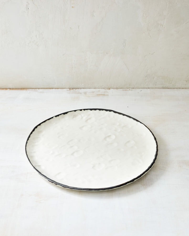 DBO HOME Pinch Dinner Plate in hand-formed white porcelain with a bronze-edged rim. Subtle indentations and an organic silhouette highlight the artisanal process, styled on a pale, textural backdrop.