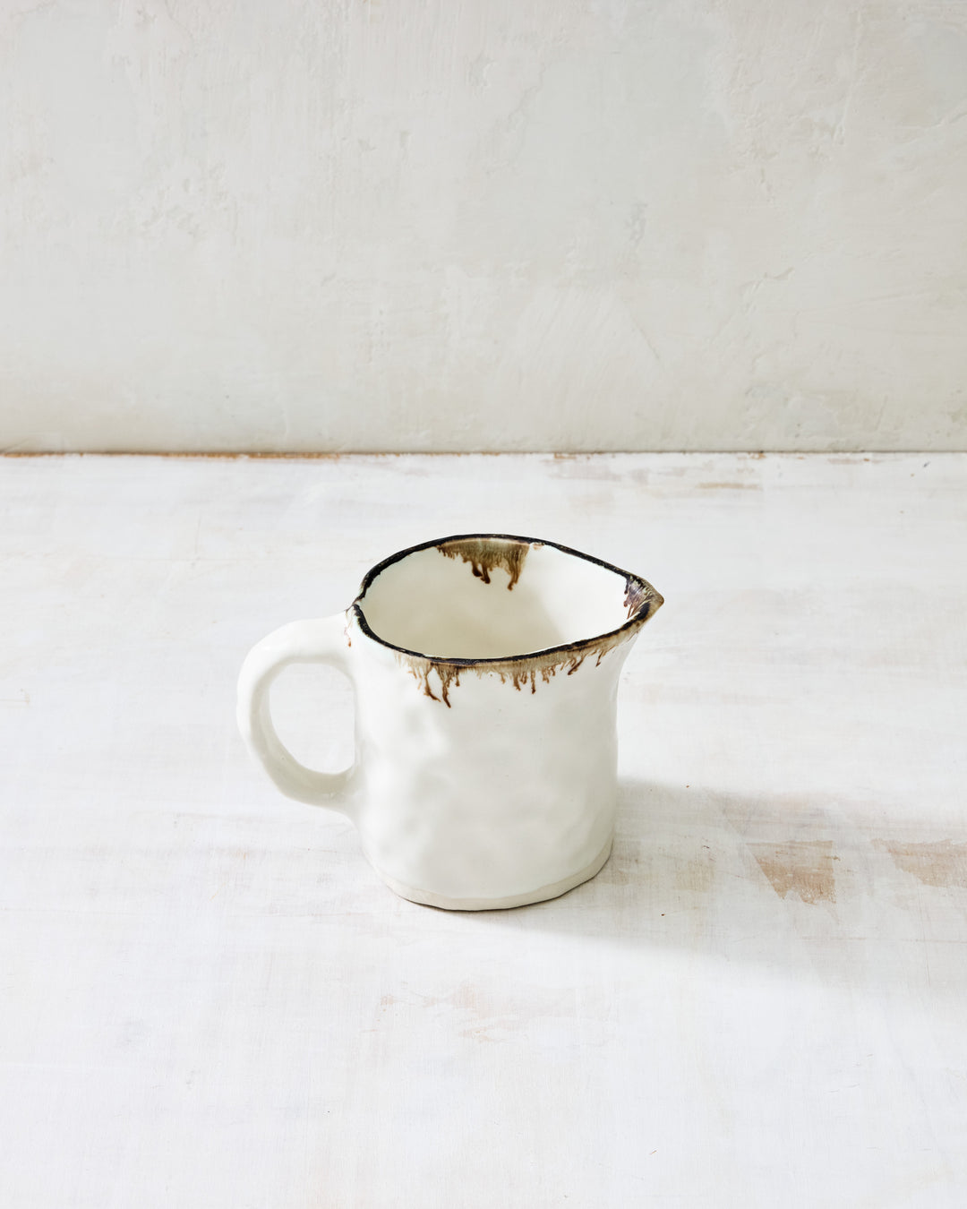 DBO HOME Pinch Jug in creamy white porcelain with a bronze-dipped rim, handcrafted with a sculptural spout and rounded handle. A small ceramic pitcher for cream, sauce, or syrup, styled on a rustic whitewashed surface.