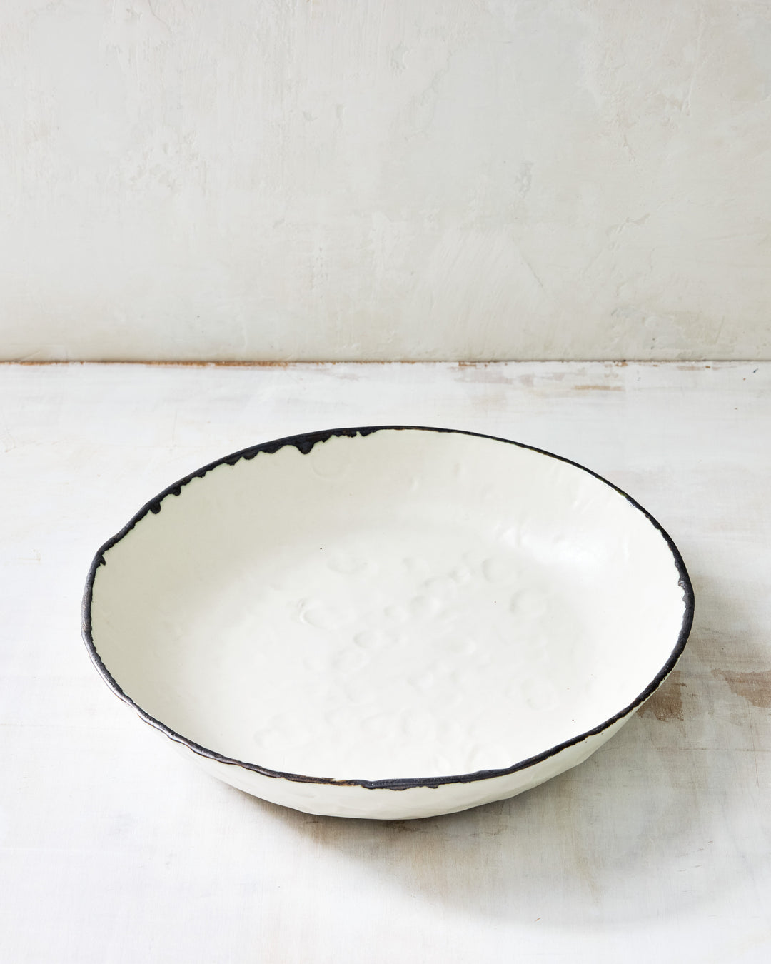 A handcrafted white ceramic serving bowl from the DBO HOME Pinch Collection, shown from an angled top-down view. The bowl features a textured surface, uneven rim, and dark glazed edge, resting on a rustic whitewashed wooden table.