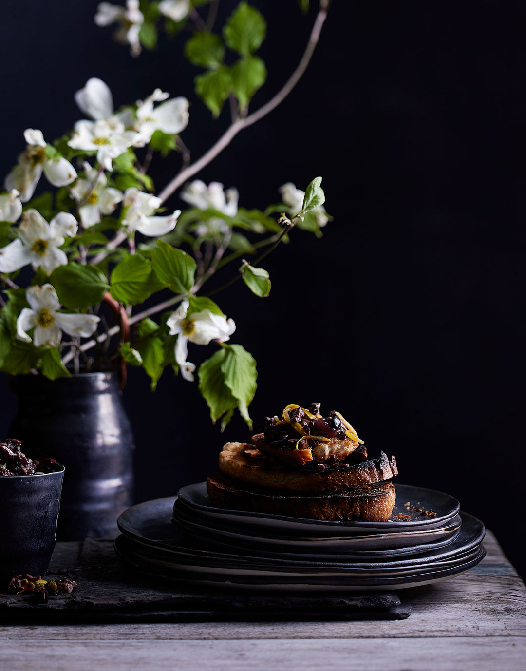 DBO HOME dramatic stack of black ceramic dinner plates and salad plates piled with food on a dark background with bright white flowers and green leaves