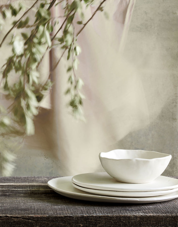 DBO HOME handmade porcelain cream colored Bare Soup Bowl and stack of artisan ceramic plates on natural wood table with softs linens and plant branches in the background