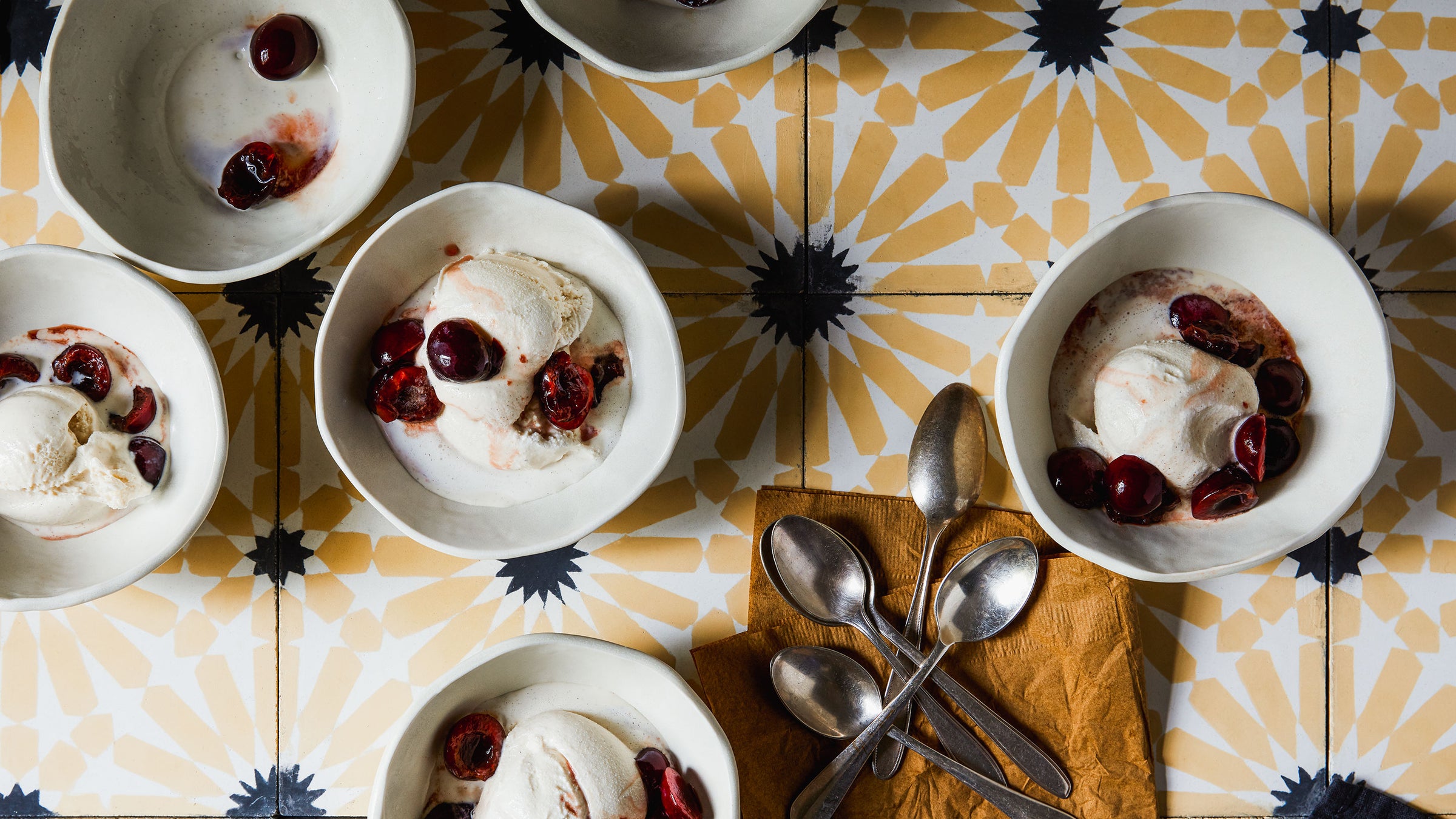 DBO HOME handmade white ceramic bowls with vanilla ice cream and ruby red cherries on yellow and blue flower tiles and a pile of spoons