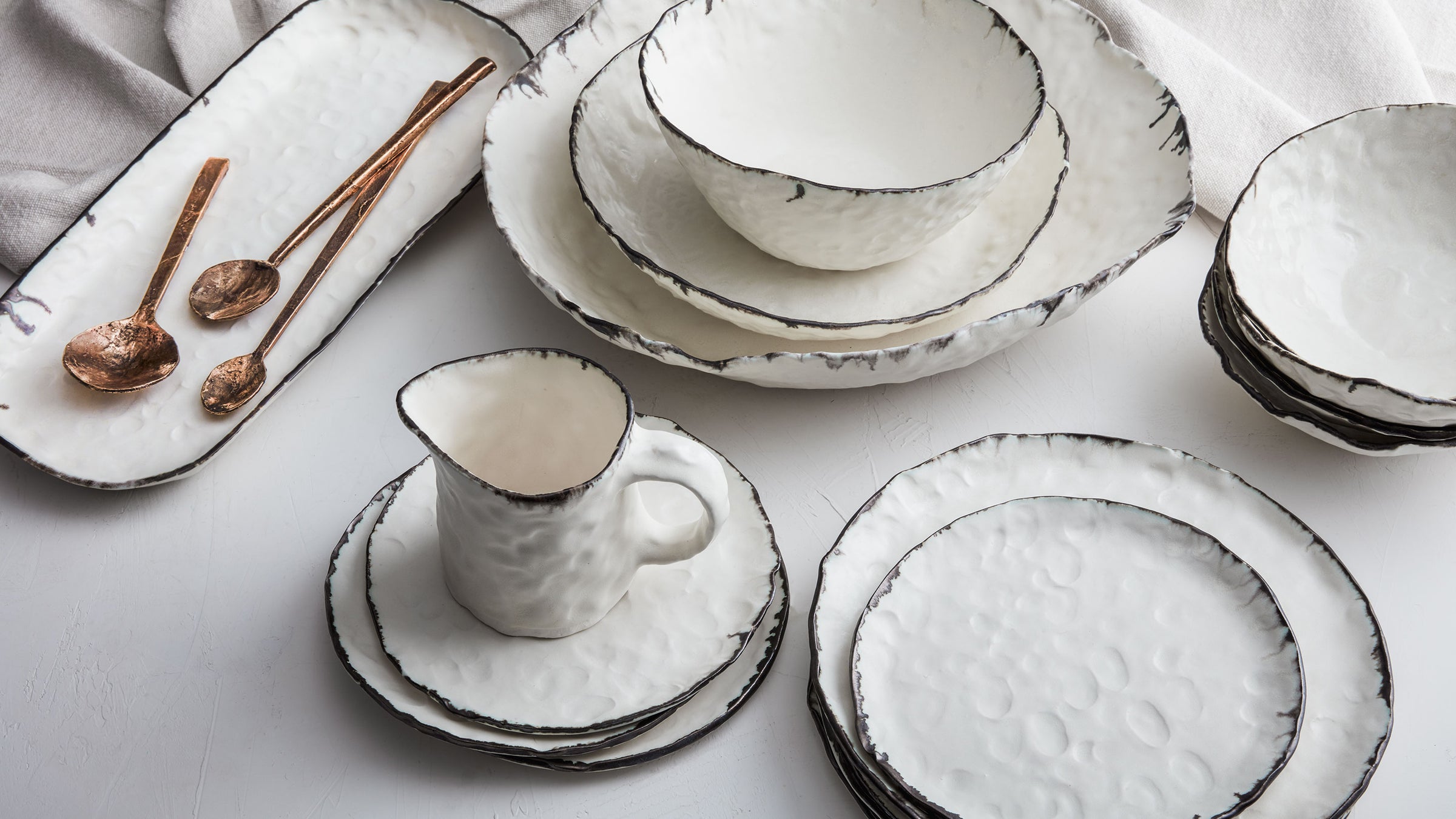 dbo home artisan porcelain textured pinch plates, bowls, trays and pitcher on a white linen tablecloth with bronze cast serving spoons