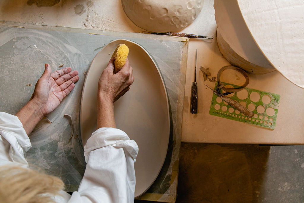 DBO HOME Artisan Handmade Luxury Ceramics | Birdseye view of Dana Brandwein in her Litchfield Country Connecticut studio using a sponge to smooth an in-process oval serveware piece, surrounded by tools and work materials