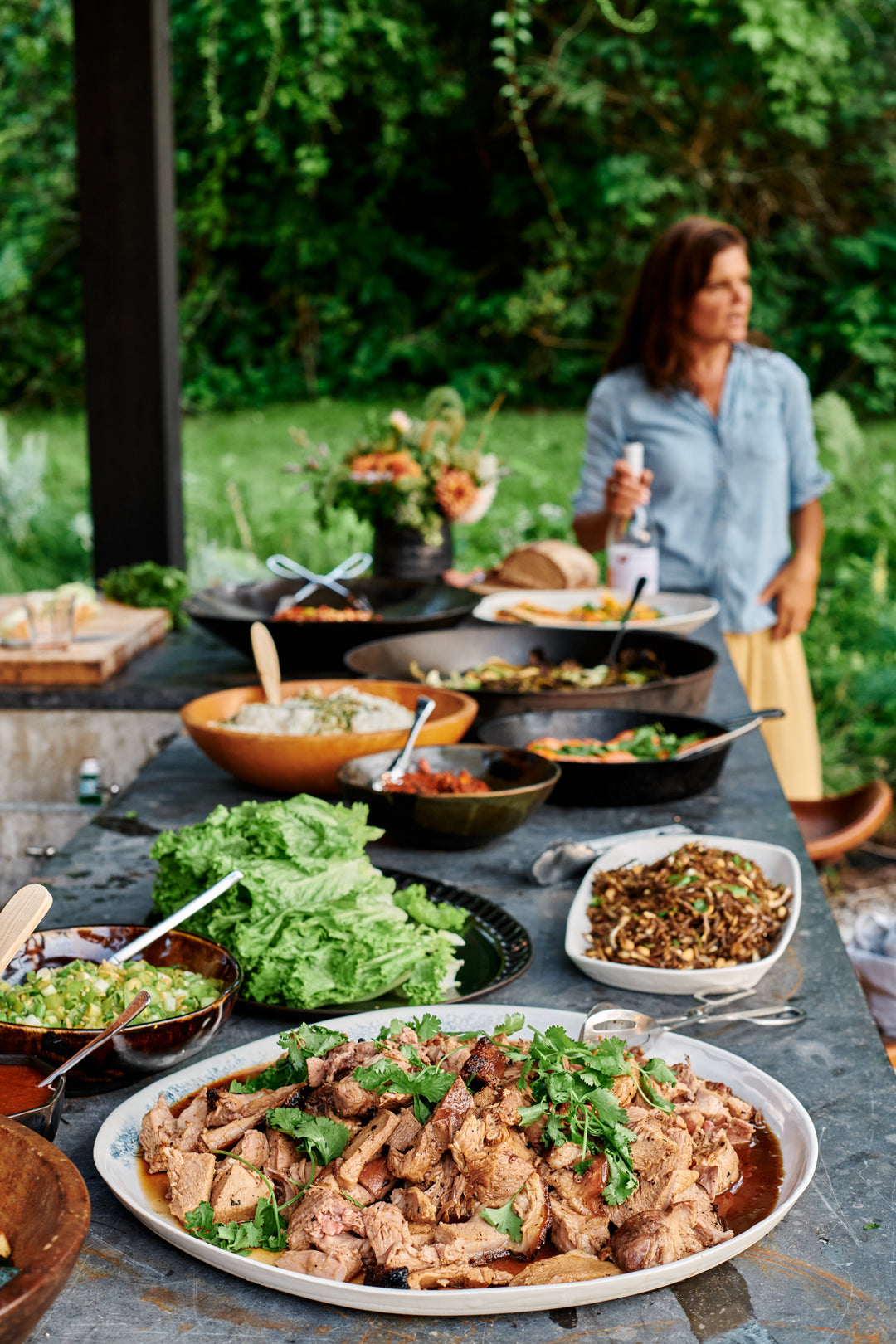 Outdoor buffet on a dark stone counter with sliced pork and cilantro, lettuce leaves, noodles, rice and sauces, a woman holding a bottle in the background. Includes the Nonna Oval Platter in Matte Black, Kashmir Large Round Bowl and Bare Long Tray in Snowflake.