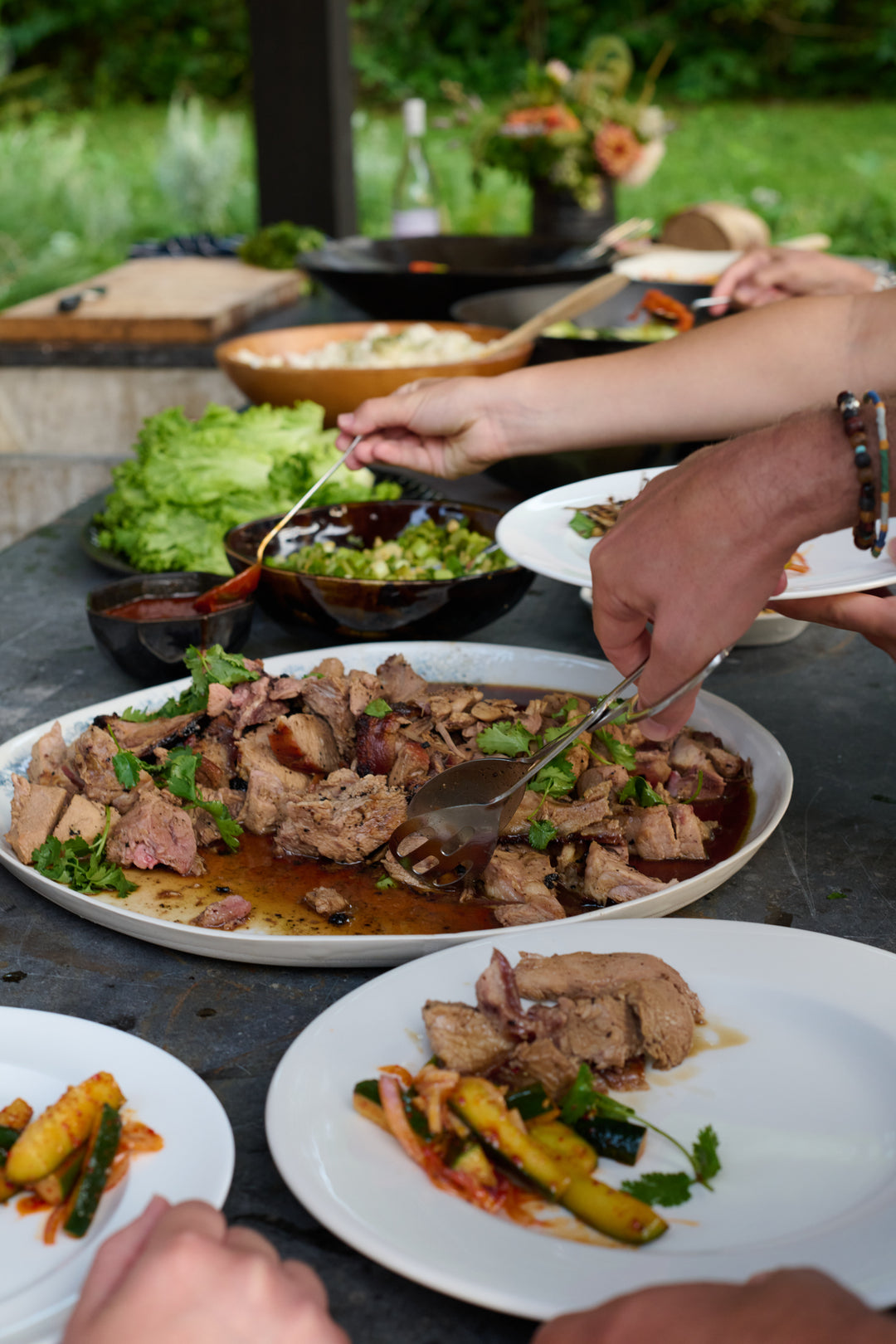 Guests serving themselves sliced pork with tongs from a large white platter, beside scallion relish in a brown bowl, lettuce leaves and chili sauce, with plates of spicy cucumber salad in front. Includes Wabi Ramen Bowl in French Roast, Kashmir XL Platter and Battuto Dessert Bowl in Mussel.