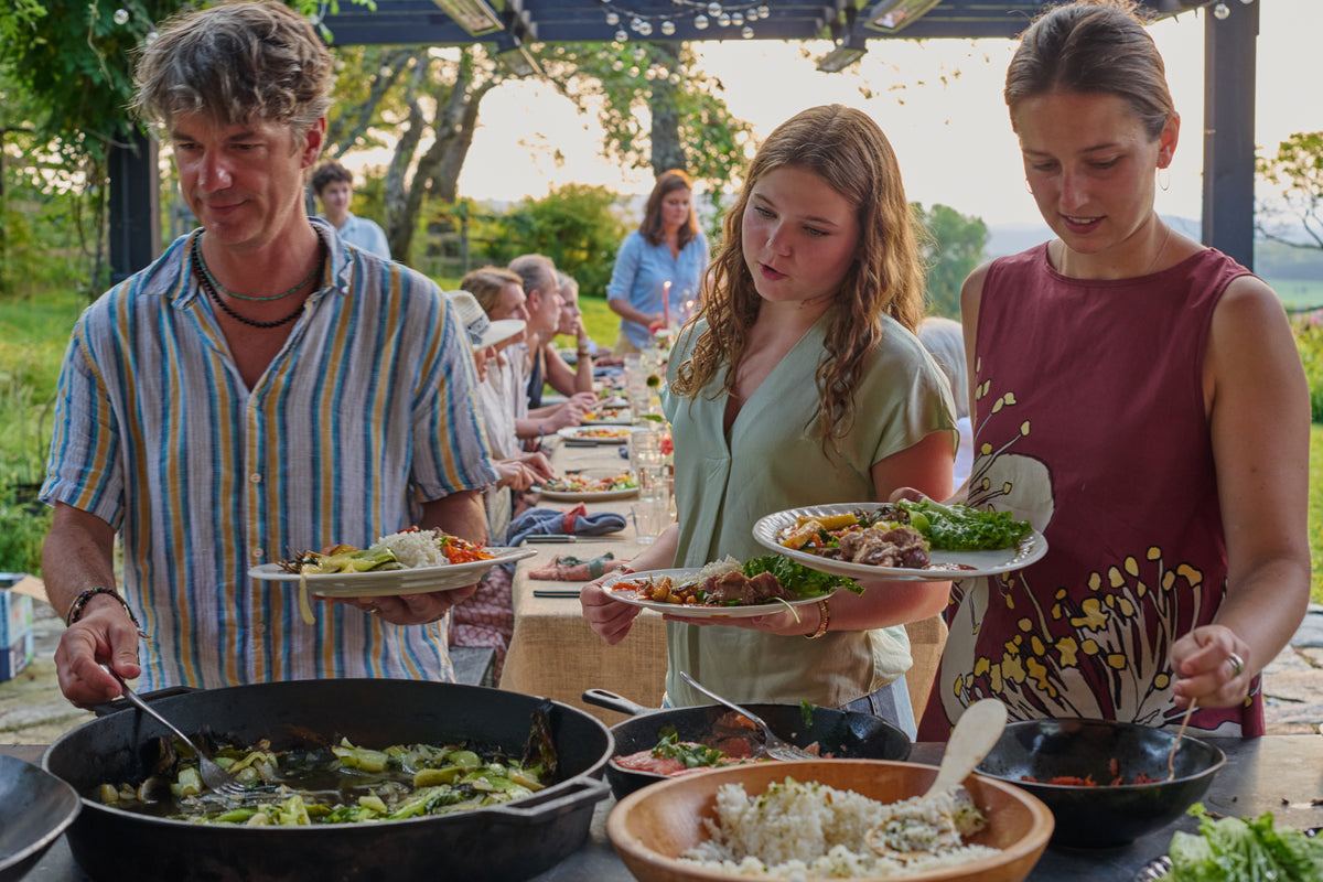 Guests filling their plates at an outdoor buffet under a pergola, with charred bok choy in a cast iron pan and rice in a wooden bowl, and a long dinner table behind them.