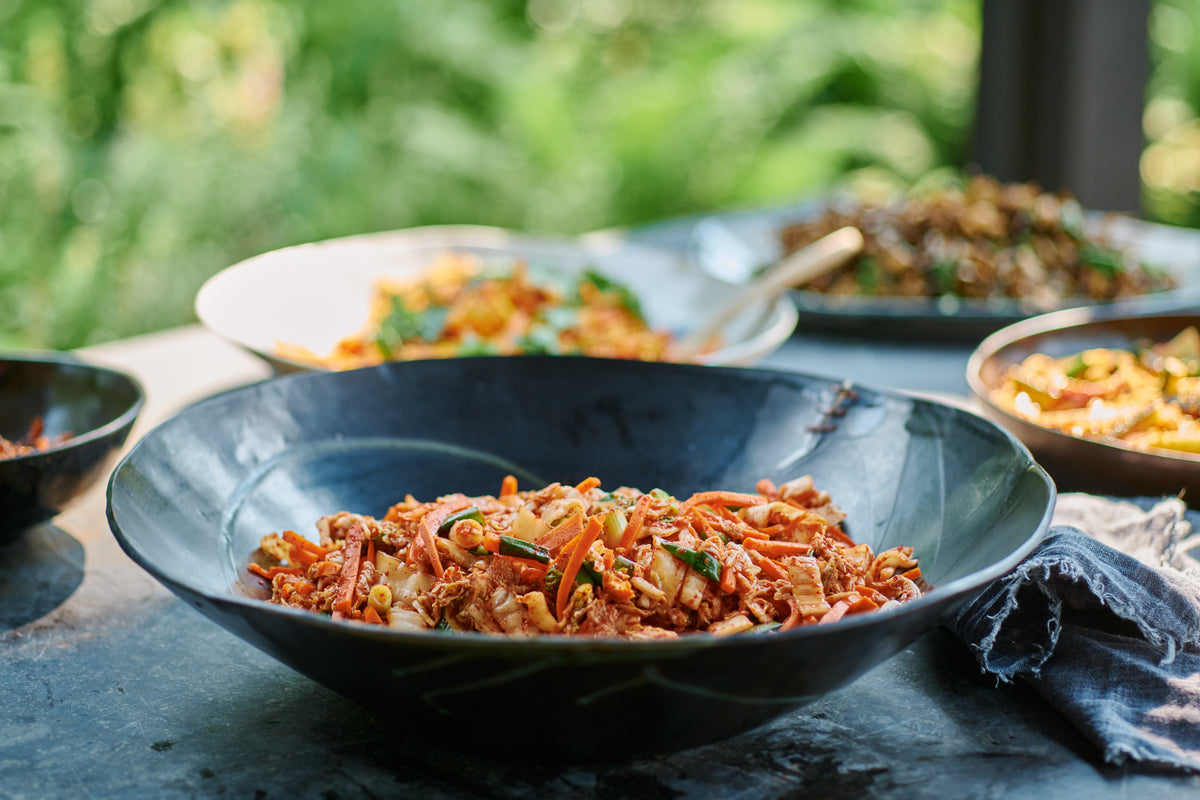Fresh kimchi with carrot and scallion in a wide, dark one-of-a-kind DBO Home bowl, with more dishes and a garden blurred behind.