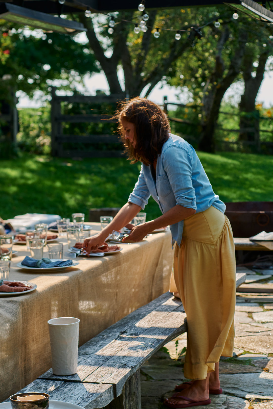 A woman in a chambray shirt and yellow skirt setting an outdoor table with plates, linen napkins and glassware, with a white Stucco Slim Vase on the wooden bench in front.