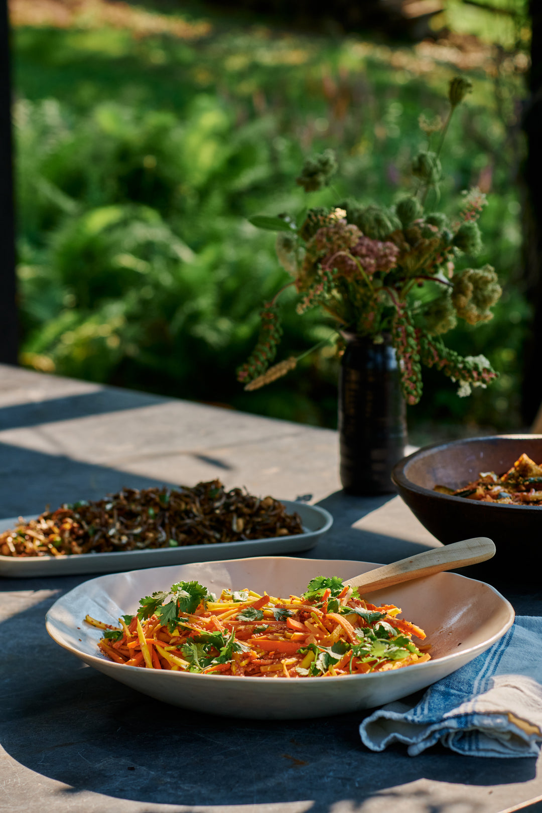 Carrot salad with cilantro and a wooden spoon in a white bowl, crispy anchovies on a long tray, and a wildflower arrangement in a dark vase, garden behind. Includes Bare Long Tray in Snowflake and a one-of-a-kind DBO Home piece.
