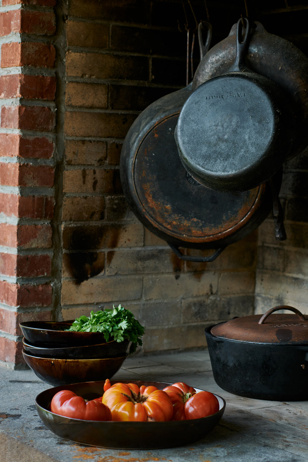 Heirloom tomatoes in a dark shallow bowl and stacked brown bowls with cilantro below cast iron pans hanging in a brick hearth. Includes Bare Large Round Bowl in Mussel and Wabi Ramen Bowl in French Roast.