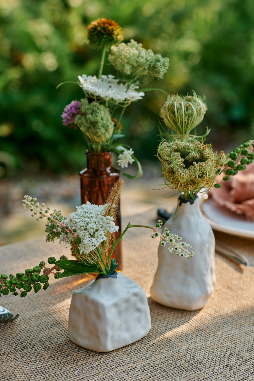 Queen Anne's lace, wildflowers and pokeweed in white textured bud vases and an amber bottle on a burlap tablecloth. Includes Pinch Bud Vase - Ringo and Pinch Bud Vase - John.
