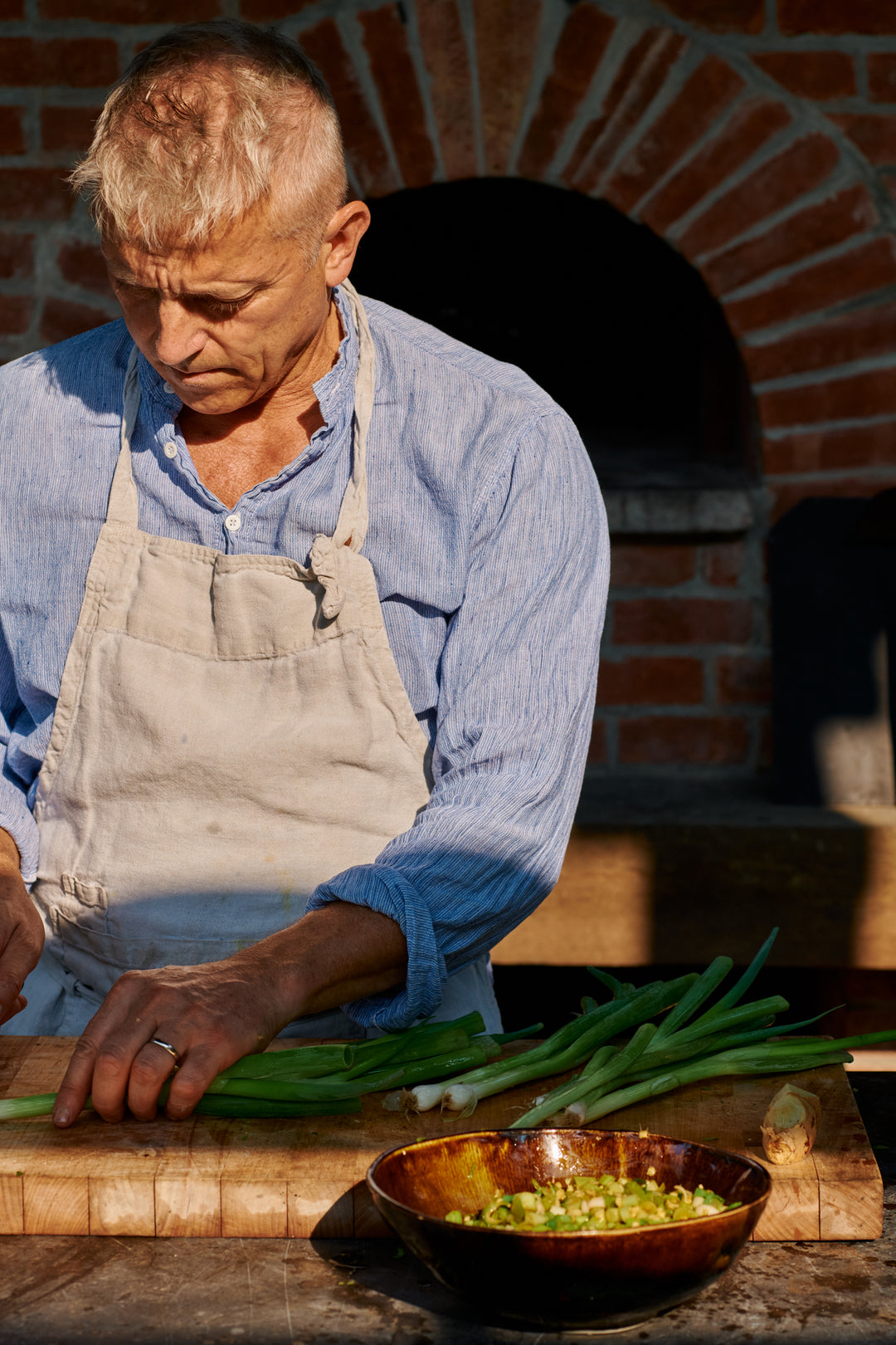 A man in a linen apron and blue shirt trimming scallions on a butcher block in front of a brick oven, with chopped scallion in a Wabi Ramen Bowl in French Roast.