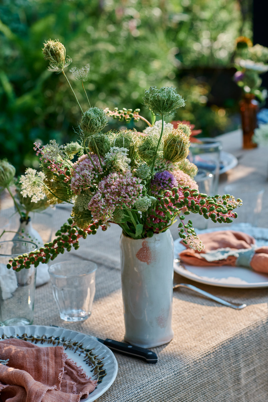 Wildflower arrangement of Queen Anne's lace, sedum, allium and pokeweed in a one-of-a-kind white stamped DBO Home vase, on a burlap-covered table set with Nonna Dinner Plates in Bronze Drip and pink linen napkins.