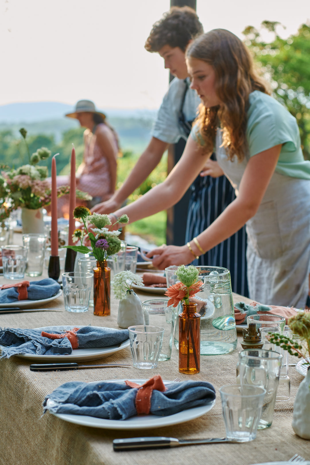 Two young people in aprons setting a long outdoor table with blue linen napkins, glassware, amber bottles of flowers and pink taper candles, with hills behind.