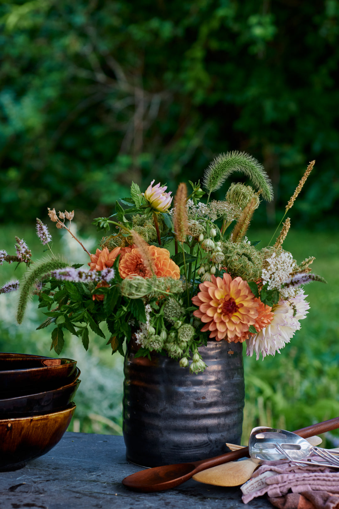 Arrangement of peach dahlias, mint, Queen Anne's lace and grasses in a dark ribbed vase, beside stacked brown bowls and wooden serving spoons. Includes Wabi Ramen Bowl in French Roast and a Mended piece.