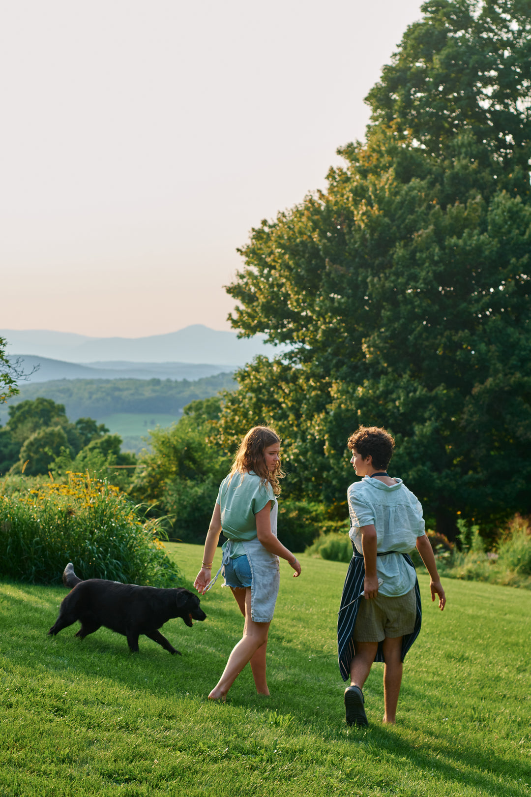 Two young people in aprons walking across a lawn with a black dog, toward a wide view of hills and mountains at dusk.