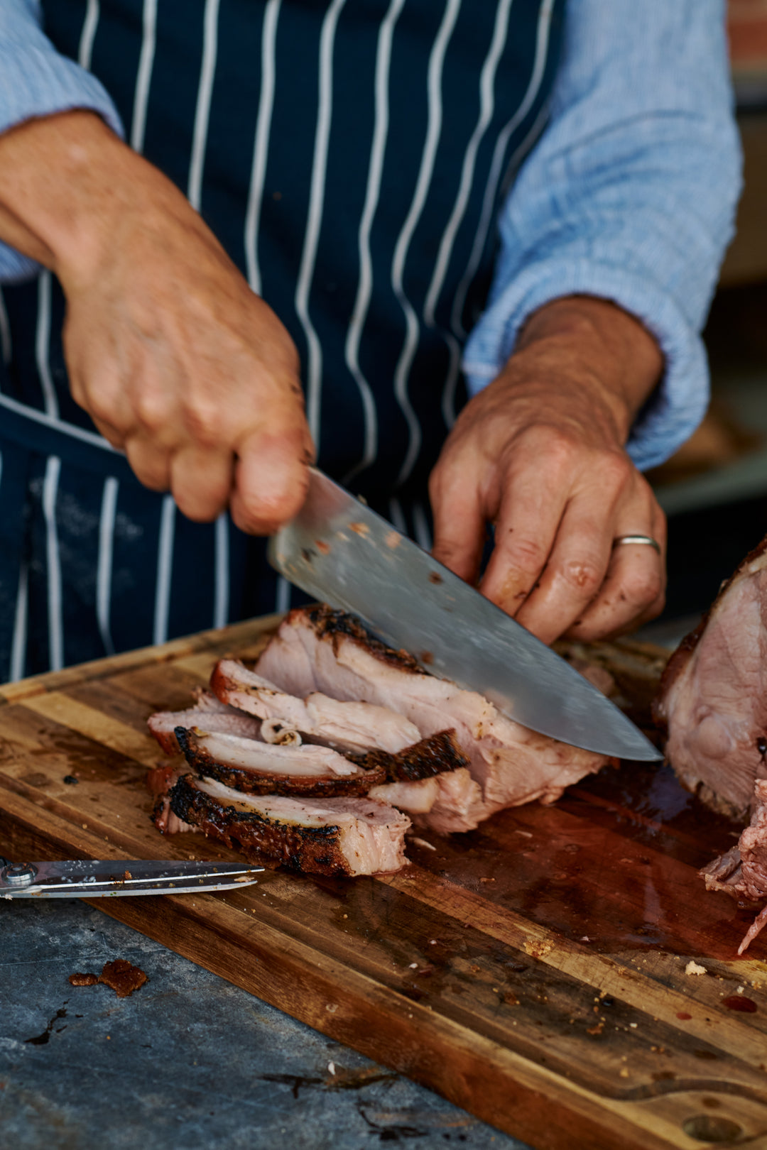 Close-up of hands in a striped apron slicing roast pork with crackled skin on a wooden cutting board.