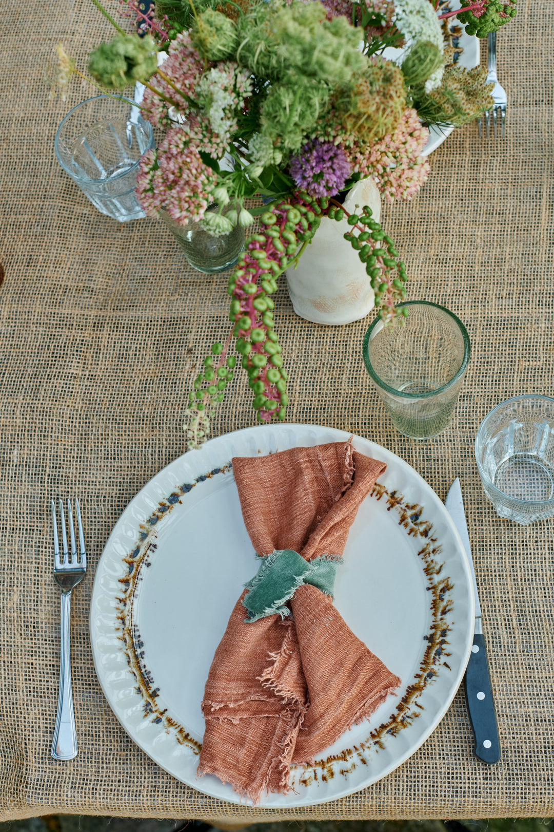 Overhead view of a Nonna Dinner Plate in Bronze Drip with a pink linen napkin tied in green, fork and knife, beside a wildflower arrangement trailing green pokeweed berries on a burlap tablecloth.