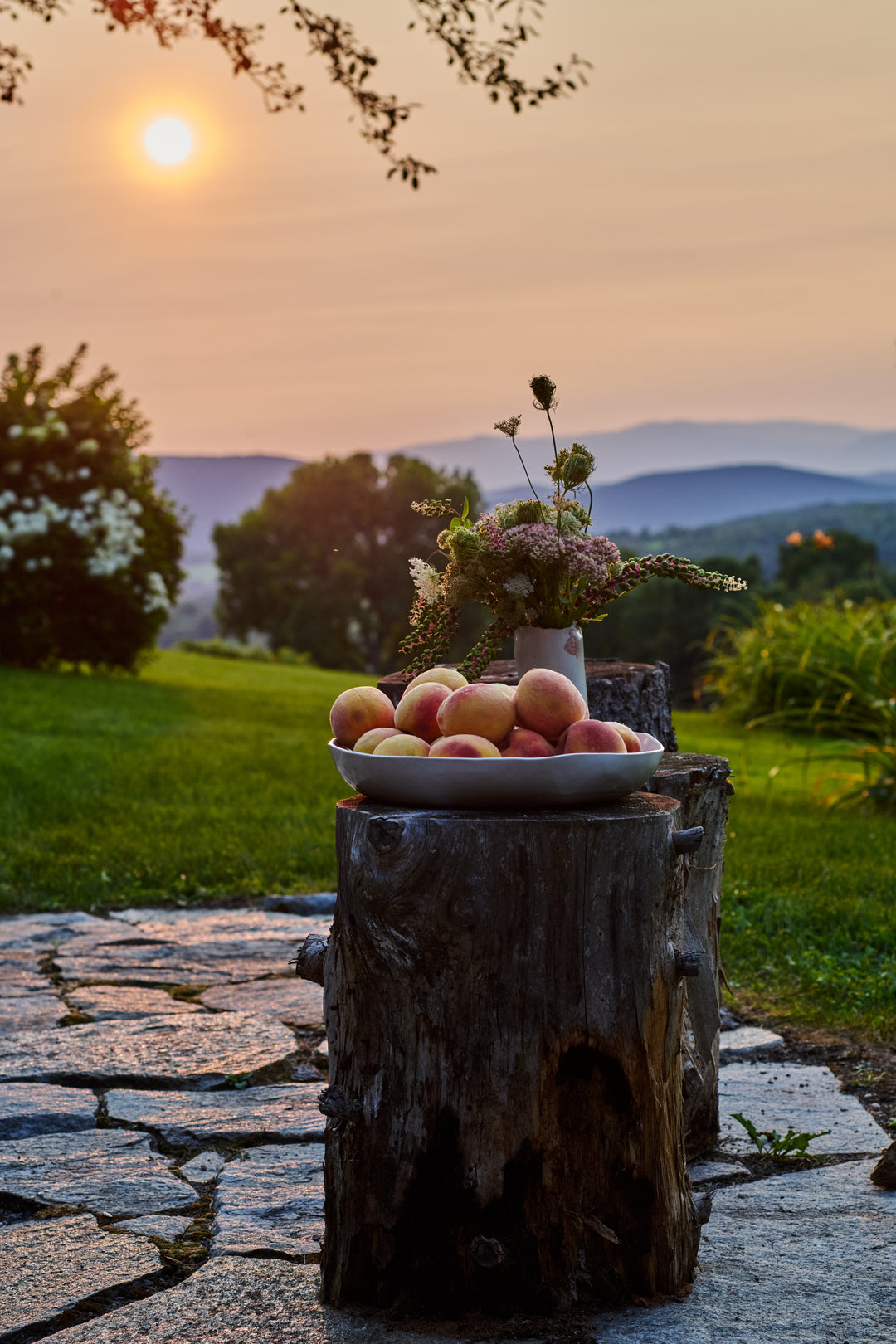 A white bowl of peaches and a wildflower arrangement in a white vase on a tree stump, with the sun setting over hazy mountains. Includes Birch Large Round Bowl.