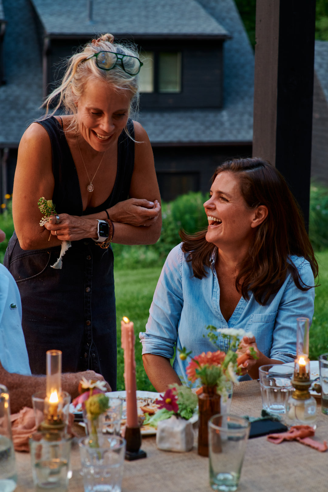 Two women laughing together at an outdoor dinner table, one standing in a black dress and one seated in a chambray shirt, with candles, flowers and oil lamps on the table.