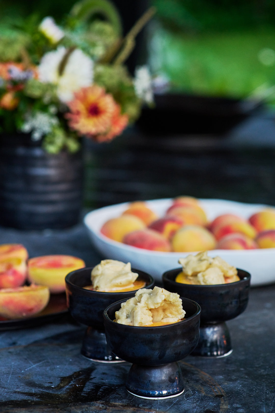 Three dark footed dessert cups of peaches topped with ice cream, with halved peaches on a dark plate, a white bowl of whole peaches and a flower arrangement behind. Includes Birch Large Round Bowl and Bare Coupe in Mussel.