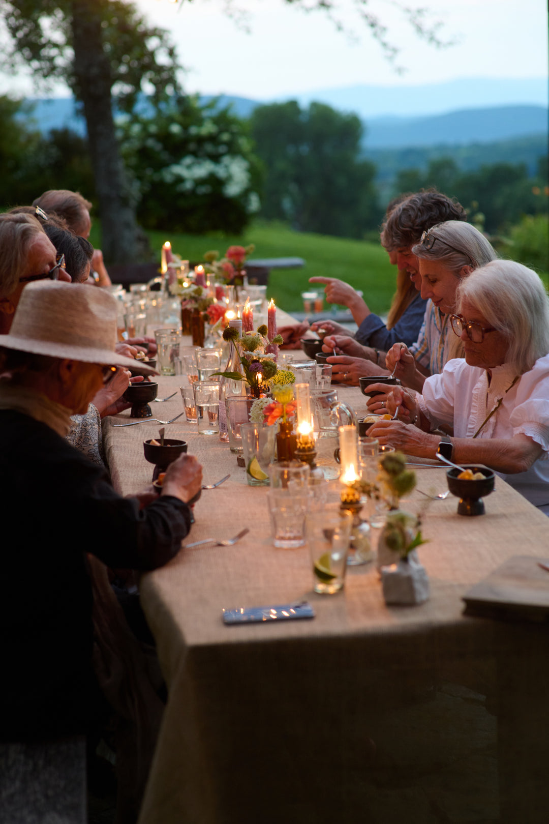 Guests eating dessert from small dark footed bowls at a long candlelit table at dusk, with a wide mountain view behind.