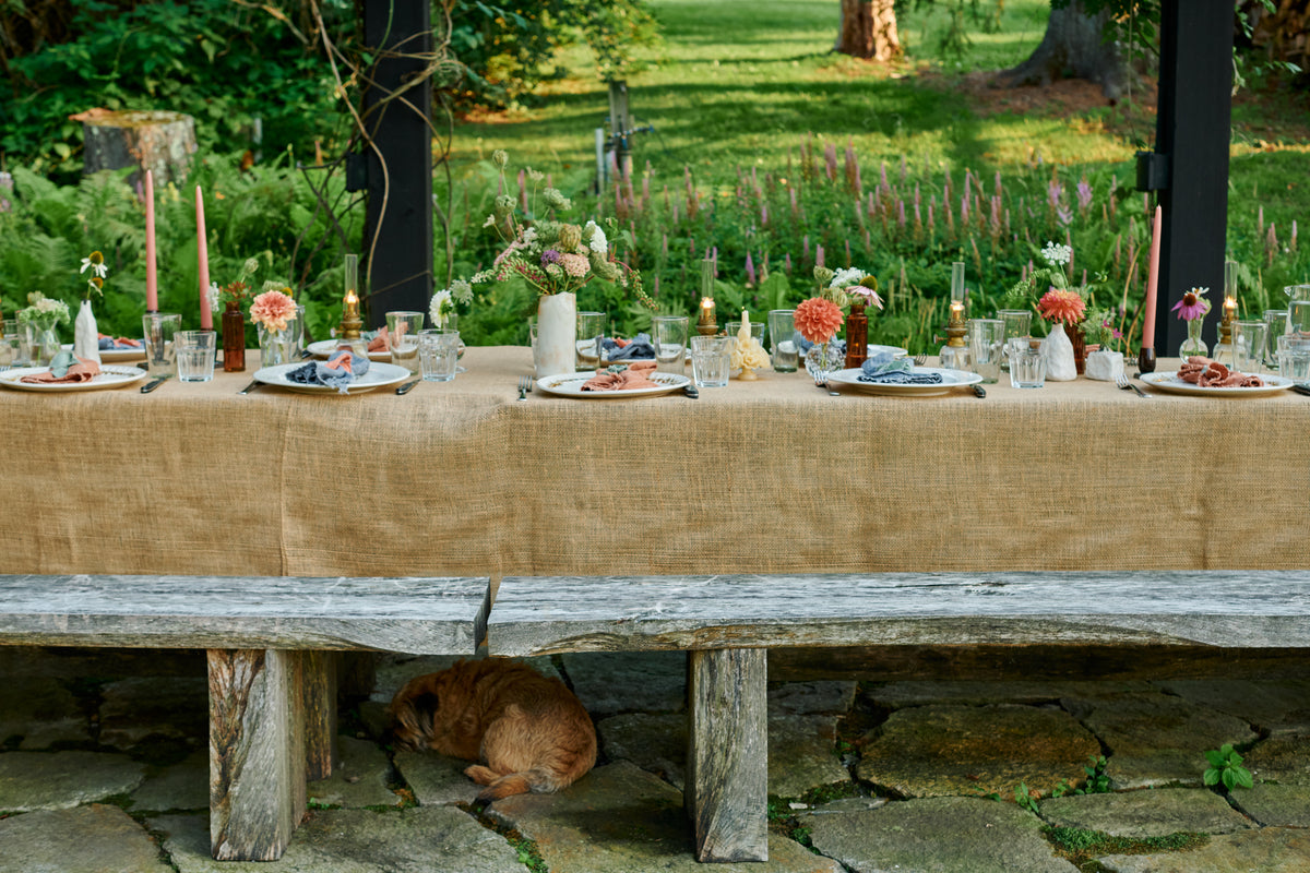 Long outdoor dinner table set with Nonna Dinner Plates in Bronze Drip, linen napkins, pink taper candles, brass oil lamps and bud vases of summer flowers, with a dog curled up asleep under the weathered wood bench.