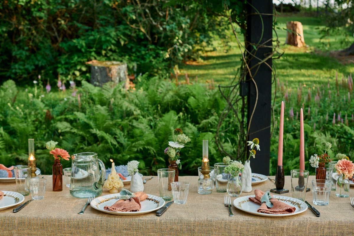 Outdoor table set with Nonna Dinner Plates in Bronze Drip and pink linen napkins, lit beeswax candles and brass oil lamps, pink tapers, a glass pitcher, and single stems in amber bottles and white bud vases, ferns behind.
