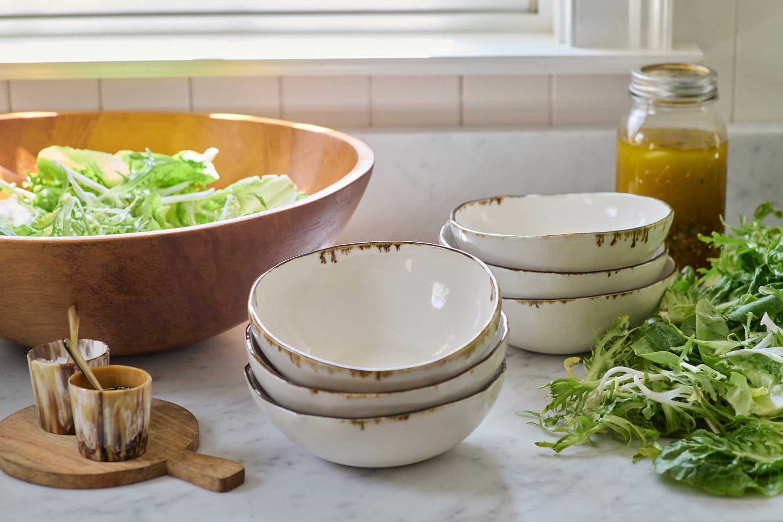 Two stacks of DBO HOME bronze drip bowls on a sunny marble kitchen counter with a big wooden salad bowl and salad greens piled on the table, plus small decorative salt and pepper dishes and a mason jar of homemade salad dressing.