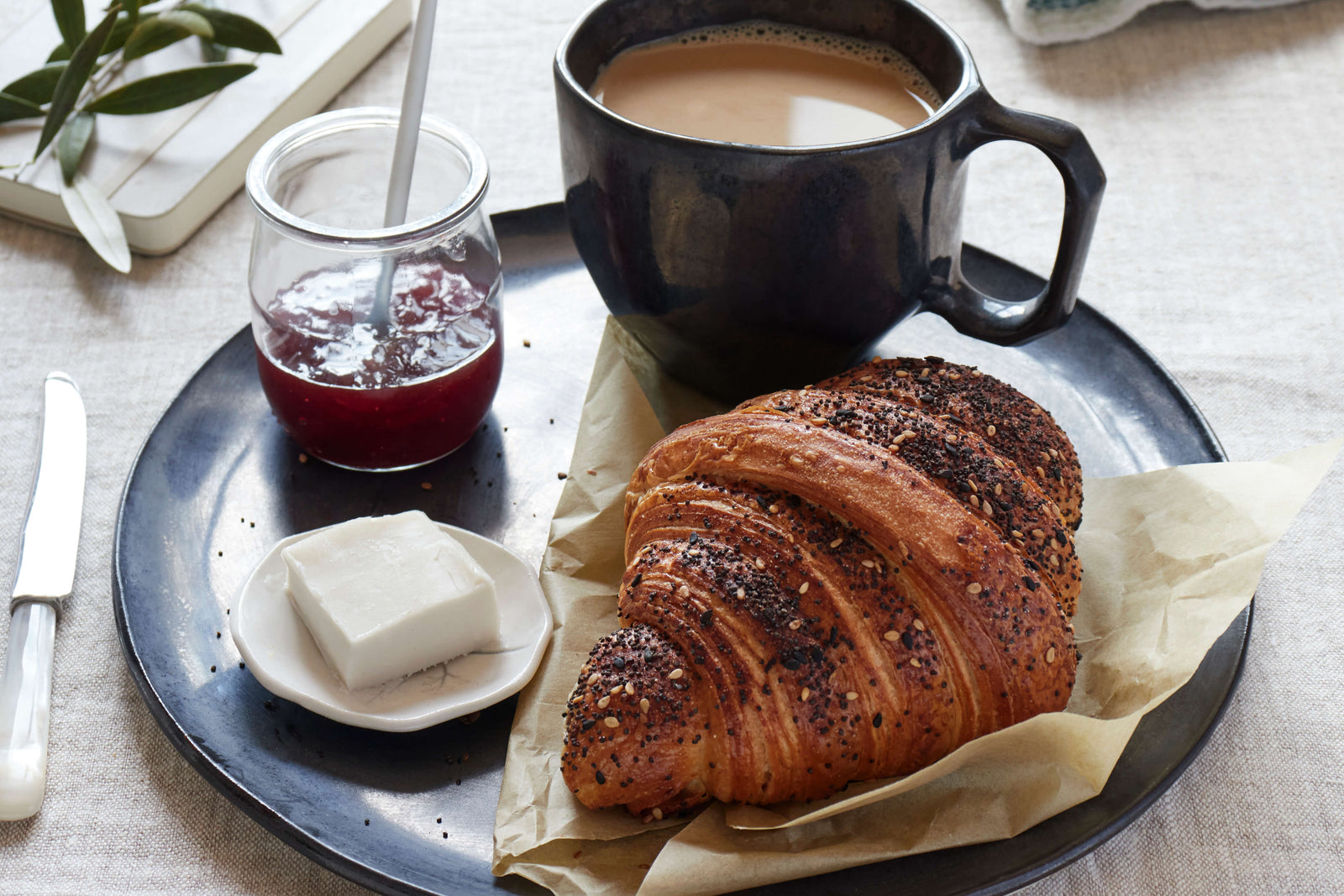 Croissant and Jam from Bourke Street Bakery on a DBO Home Bare Salad Bowl in Mussel along with a DBO Home Battuto Mug filled with coffee and a vintage DBO dipping bowl with cream cheese on a brightly lit, linen covered table