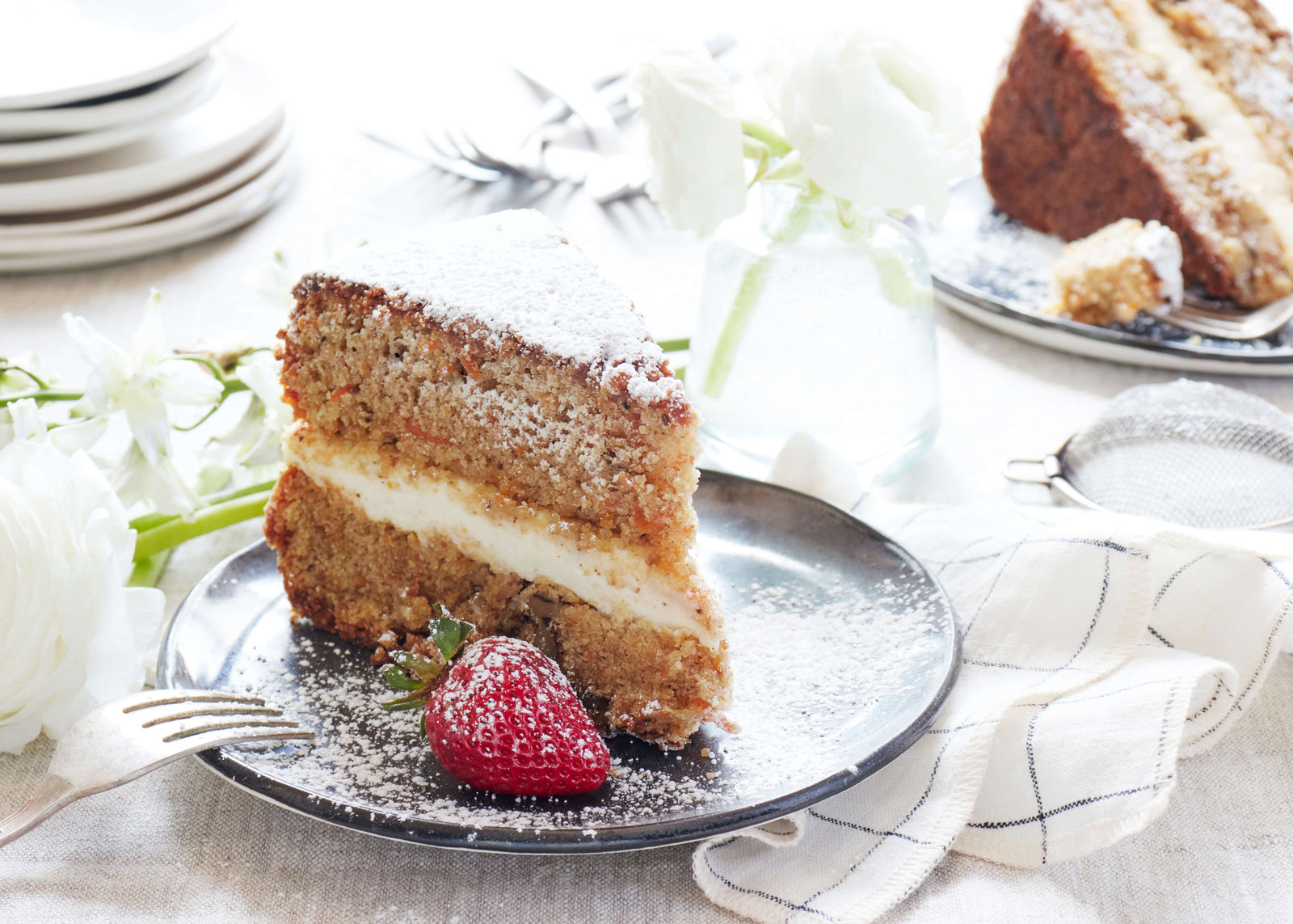 Slice of layer cake from Bourke Street Bakery dusted with powdered sugar on a DBO Home Bare Bread Plate in Mussel, on a brightly lit linen-covered table with white napkins and flowers