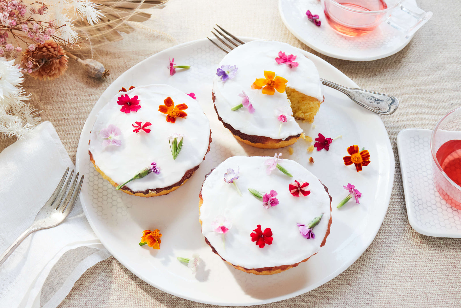Three Orange & Almond Cupcakes from Bourke Street Bakery with white frosting, covered in bright and colorful tiny flowers on a DBO Home Honeycomb Dinner Plate, with colorful drinks in vintage glasses resting on DBO Honeycomb Serveware