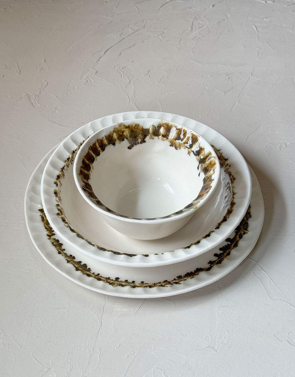 Three distinctive coordinating ceramic pieces from the DBO HOME Nonna Collection - a dinner plate, pasta bowl, and small bowl - sit stacked on a plaster surface. Each piece is an off-white natural white with a delicate and distinctive fluted rim, adorned with organic and unique bronze glaze drips and splashes in a delicate ring at the base of the rim.