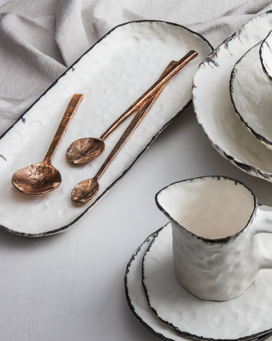 DBO HOME Pinch Long Tray in white porcelain with a bronze rim, styled with hand-forged bronze serving spoons. Surrounding pieces from the Pinch Collection—including plates, bowls, and a jug—create a layered, tactile tablescape against a soft linen backdrop.