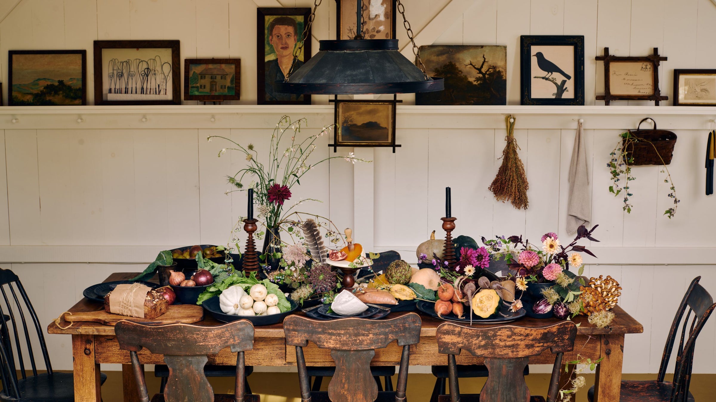 A rustic wooden table set for fall from a recent Tastemakers' Table story photographed by John Gruen, styled with DBO Home ceramics, seasonal vegetables, dried florals, and taper candles, beneath a vintage industrial pendant light and a gallery wall of artwork and natural accents.