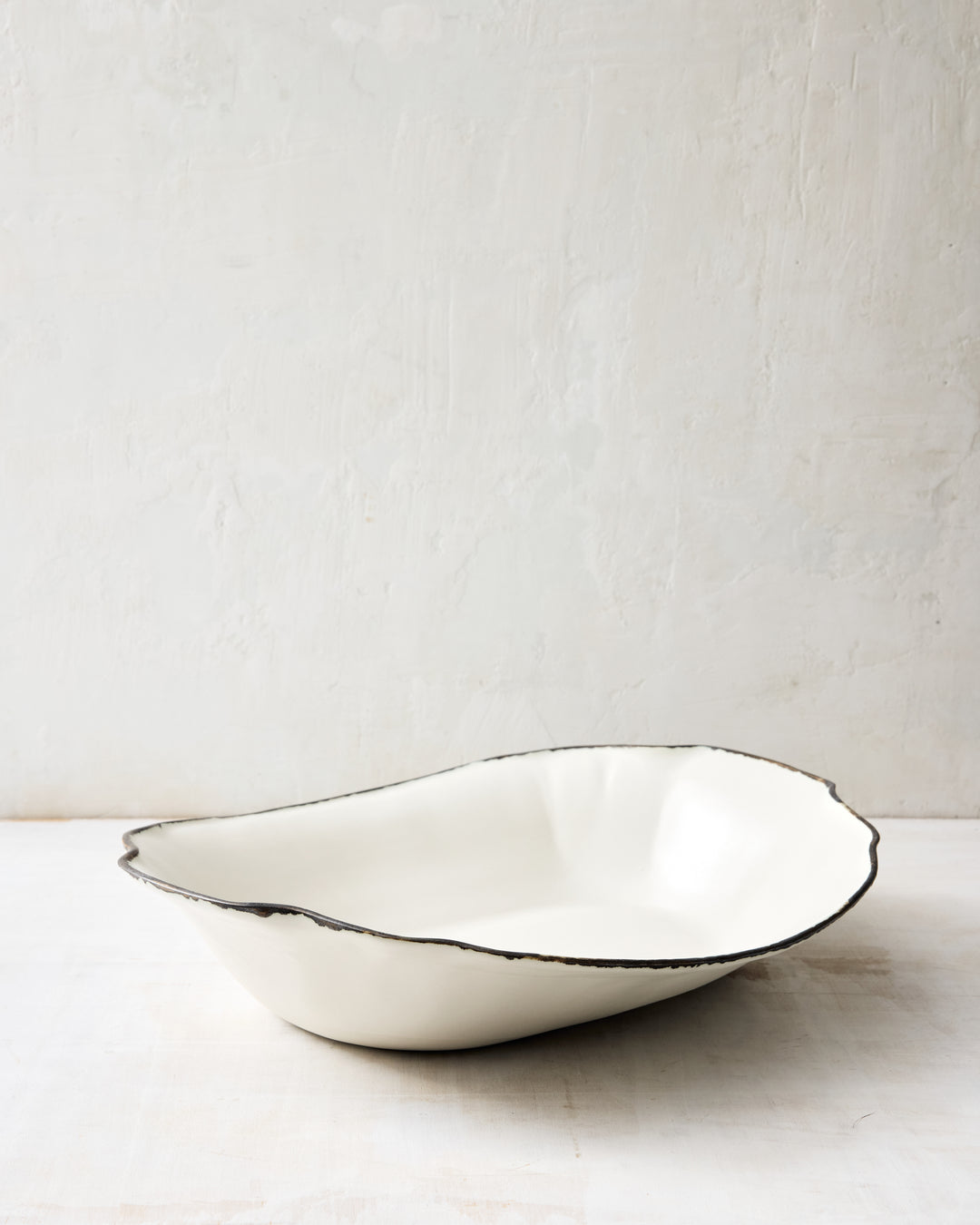 Angled DBO HOME Formale Serving Bowl in creamy porcelain with a bronze-glazed rim. The undulating edge and soft matte surface give this handmade serving bowl a grounded, elegant feel.