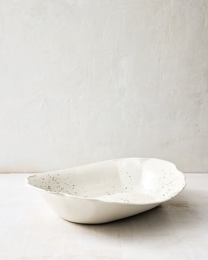Angled view of the DBO HOME Formale Serving Bowl in porcelain with painterly black speckles. Organic shaping and a wide, shallow basin make this piece ideal for entertaining or everyday meals.