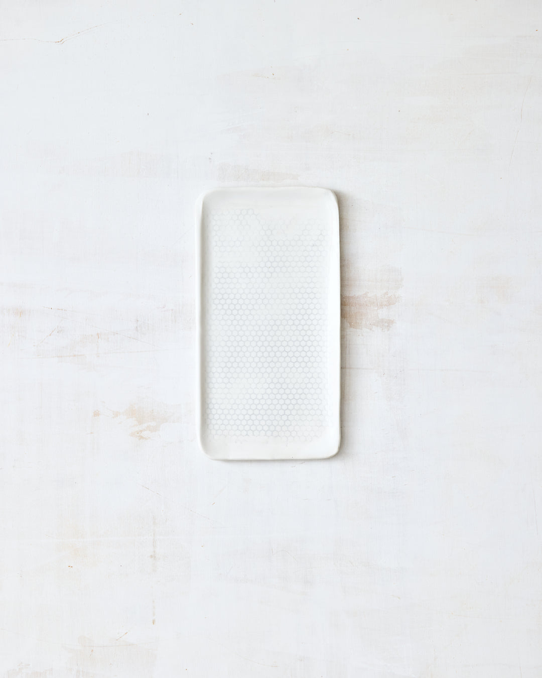 DBO HOME Honeycomb B&B Plate in handmade porcelain with a white gloss glaze and pale grey-inlaid honeycomb texture, small rectangular form with softly rounded corners, photographed overhead on a white painted wood surface.