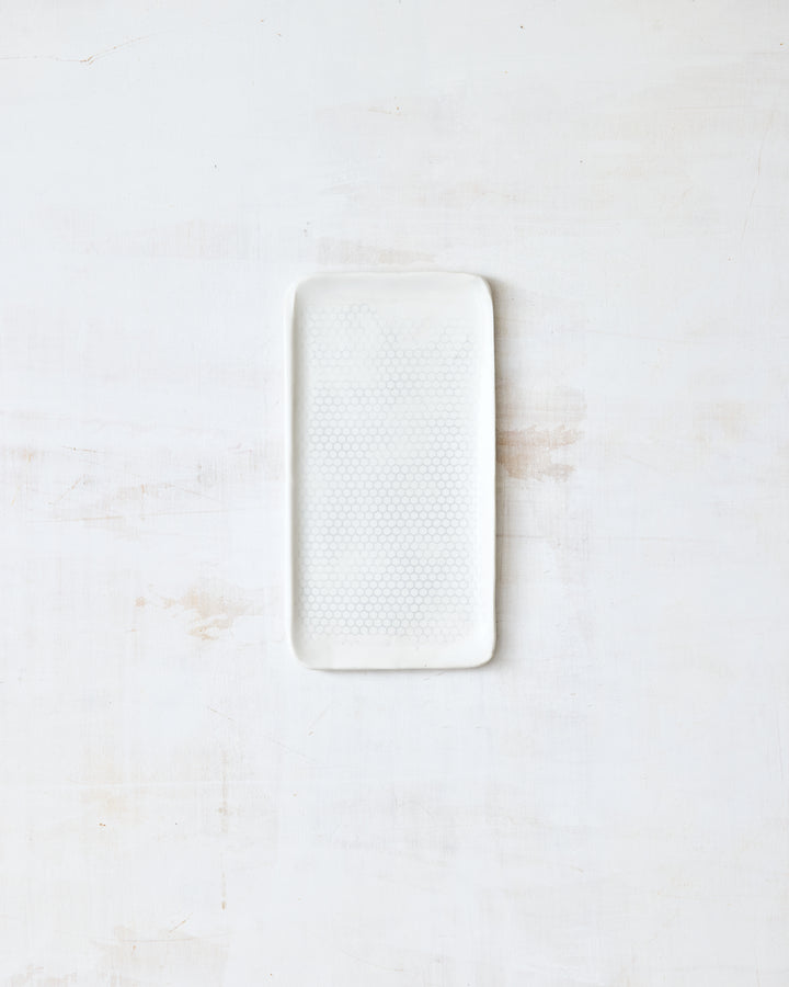 DBO HOME Honeycomb B&B Plate in handmade porcelain with a white gloss glaze and pale grey-inlaid honeycomb texture, small rectangular form with softly rounded corners, photographed overhead on a white painted wood surface.