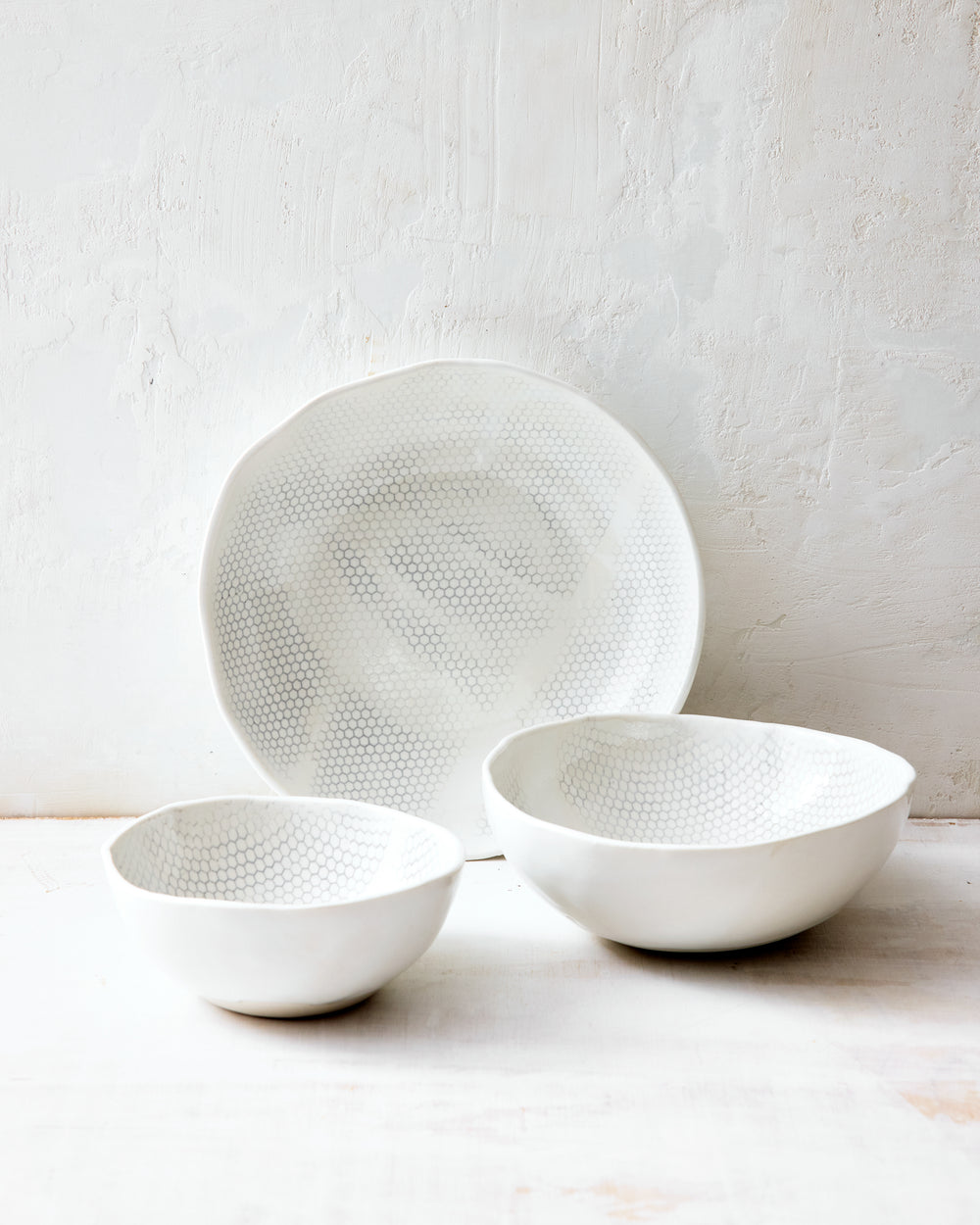 Three handmade porcelain bowls from DBO HOME's Honeycomb collection arranged together — small, medium, and large — all in a white gloss glaze with pale grey-inlaid honeycomb pattern, photographed against a whitewashed wall.