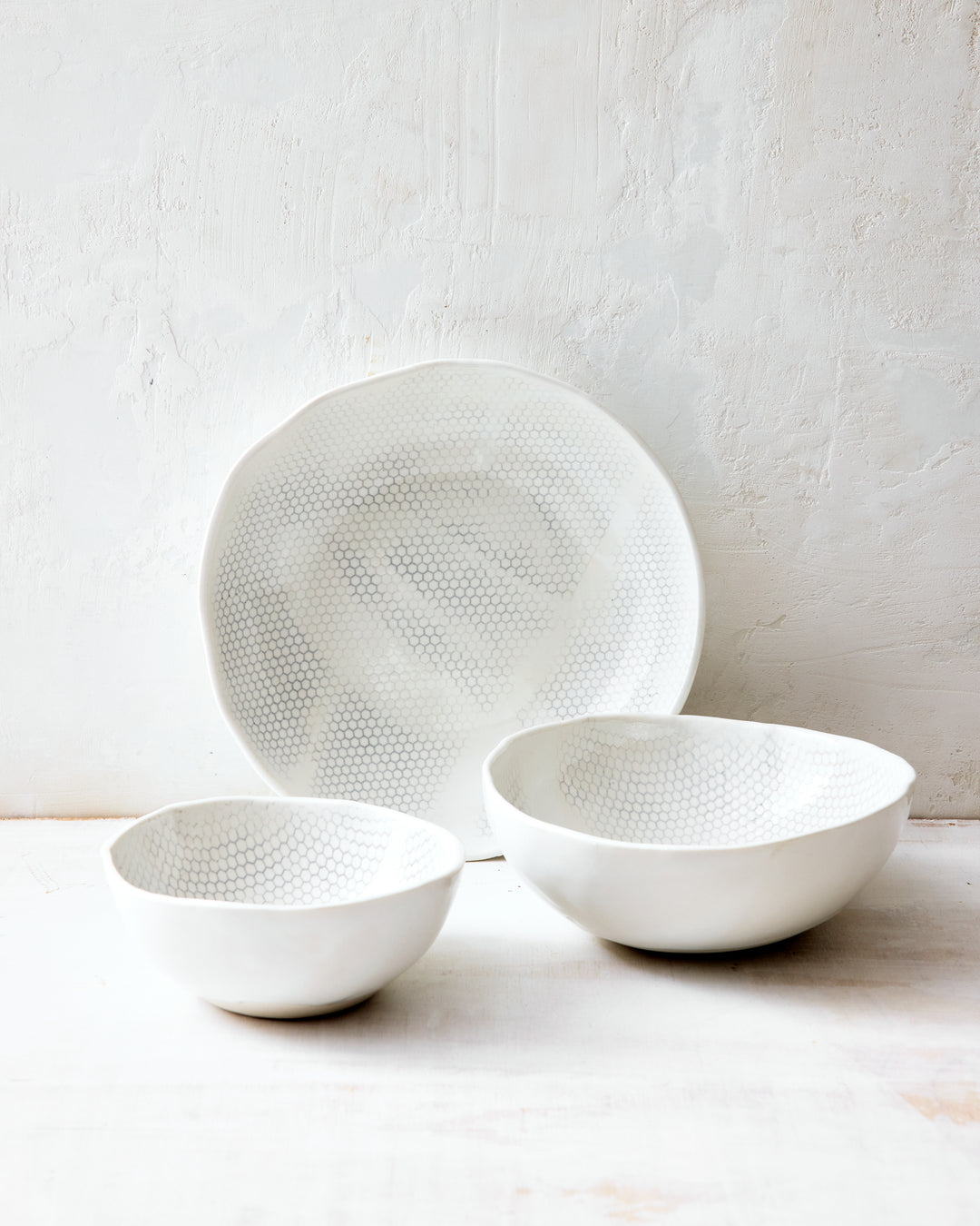Three handmade porcelain bowls from DBO HOME's Honeycomb collection arranged together — small, medium, and large — all in a white gloss glaze with pale grey-inlaid honeycomb pattern, photographed against a whitewashed wall.