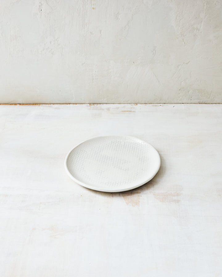 DBO HOME Honeycomb Bread Plate in handmade porcelain with a white gloss glaze and pale grey-inlaid honeycomb texture, photographed at a low angle on a whitewashed surface.